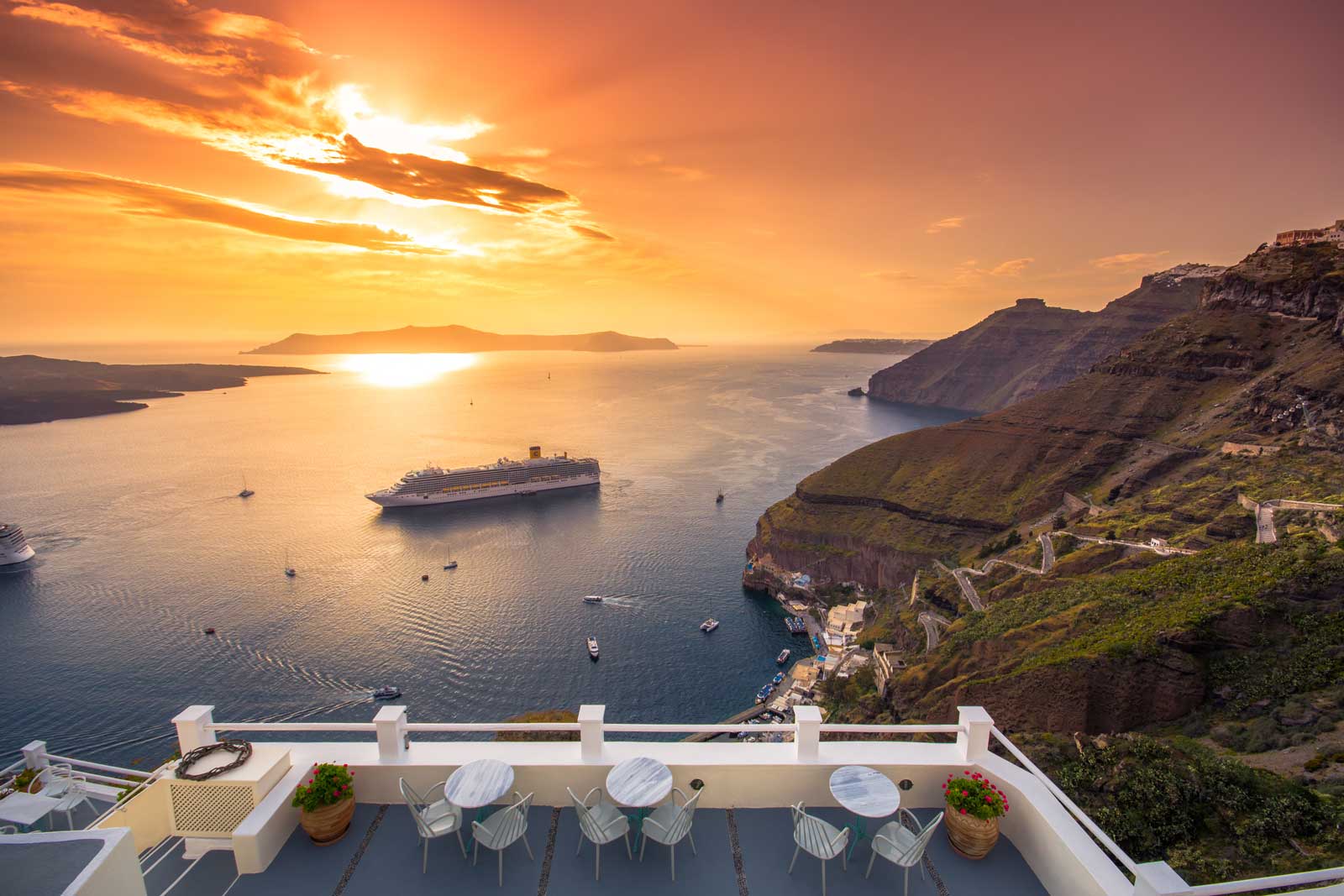 Cruise ship anchored in Santorini caldera at sunset, viewed from clifftop terrace with white railings
