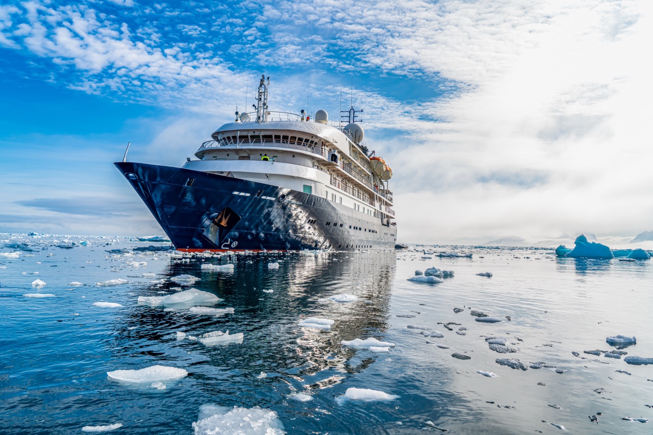 Cruise ship with blue hull surrounded by icebergs in Antarctic waters under cloudy sky