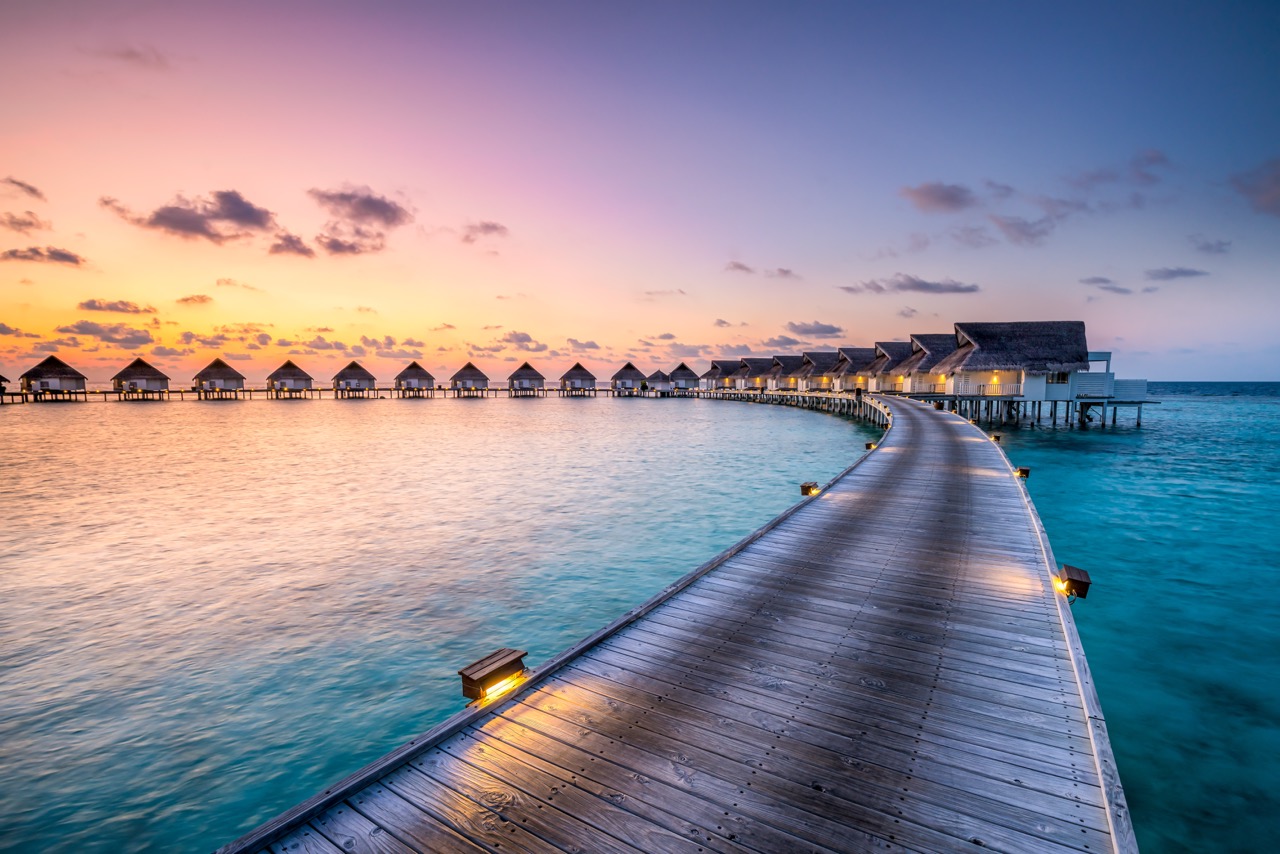 Illuminated wooden boardwalk leading to row of overwater bungalows at sunset in tropical lagoon