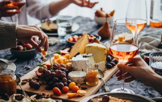 Hands reaching for cheese and grapes at a wooden board with wine glasses and charcuterie spread